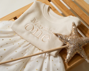 May include: A white tulle tutu skirt with gold glitter stars and moons, a white onesie with gold embroidery that says "Maya is one", and a gold star wand with sequins.
