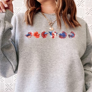 May include: A light gray sweatshirt featuring a row of colorful betta fish designs across the chest. The fish are in shades of red, blue, and white. The sweatshirt has long sleeves and a crew neck.