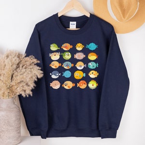 May include: Navy blue sweatshirt featuring a grid of colorful cartoon pufferfish. The pufferfish have various expressions and colors, including green, orange, blue, and yellow. The sweatshirt is hanging on a wooden hanger.