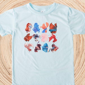 May include: A light blue t-shirt with a colorful pattern of betta fish. The fish are in various shades of red, orange, blue, and pink.
