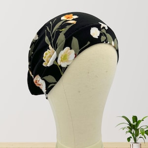May include: A black floral print hat with white, orange, and yellow flowers and green leaves. The hat is displayed on a mannequin head. The hat is a soft, comfortable material.