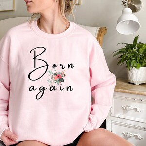 Puede incluir: Una sudadera rosa con el texto "Born again" en letra negra con un dise&ntilde;o floral.