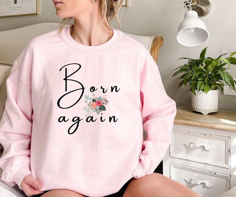 Puede incluir: Una sudadera rosa con el texto "Born again" en letra negra con un dise&ntilde;o floral.