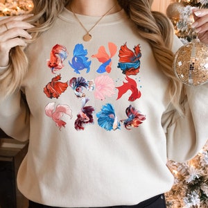 May include: A beige sweatshirt with a colorful pattern of betta fish. The fish are in various poses and colors, including red, blue, pink, and white.