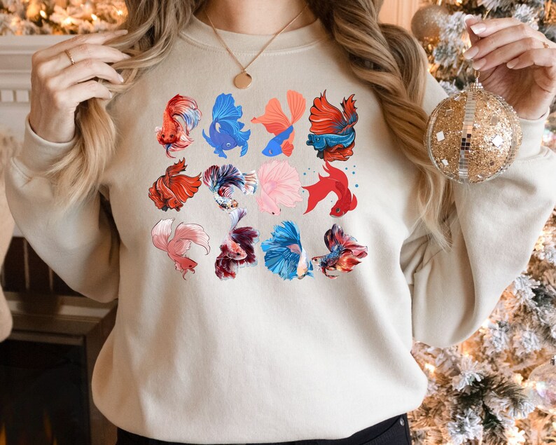 May include: A beige sweatshirt with a colorful pattern of betta fish. The fish are in various poses and colors, including red, blue, pink, and white.