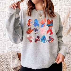 May include: A gray sweatshirt with a colorful print of betta fish. The fish are in various poses and colors, including red, blue, and orange.