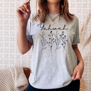 May include: Gray t-shirt with the word "Yahweh" printed in black script with a floral design below it.