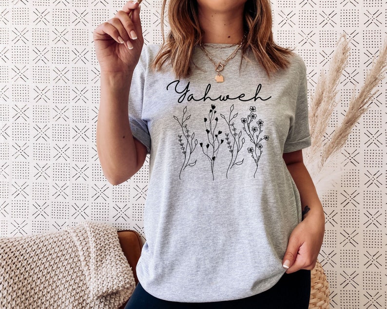 May include: Gray t-shirt with the word "Yahweh" printed in black script with a floral design below it.