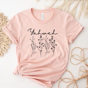 May include: A pink t-shirt with the word "Yahweh" in black script and a floral design.