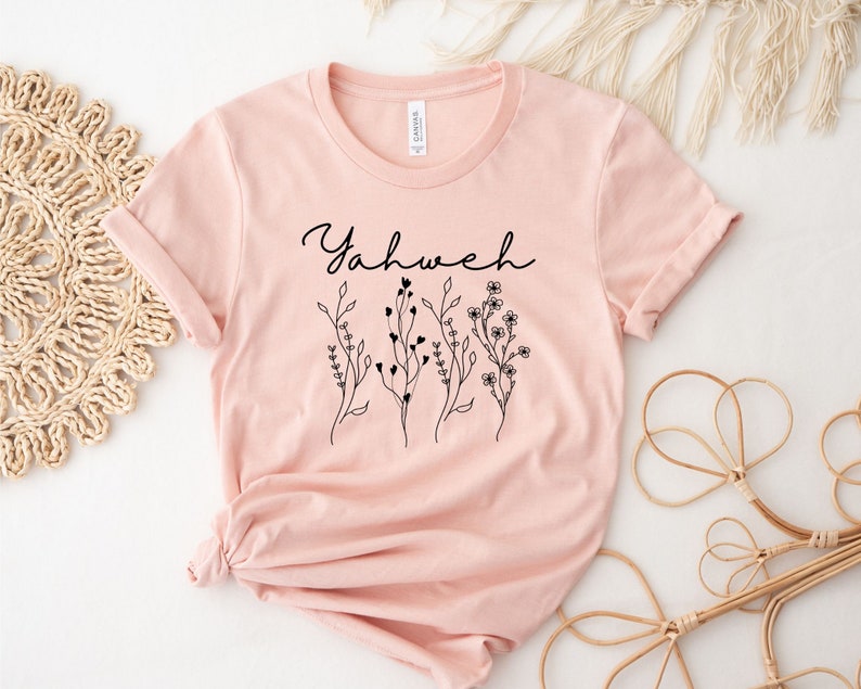 May include: A pink t-shirt with the word "Yahweh" in black script and a floral design.