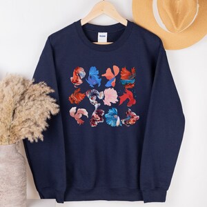 May include: A navy blue sweatshirt with a colorful pattern of betta fish. The fish are arranged in a grid pattern and are in various shades of red, orange, blue, and pink.