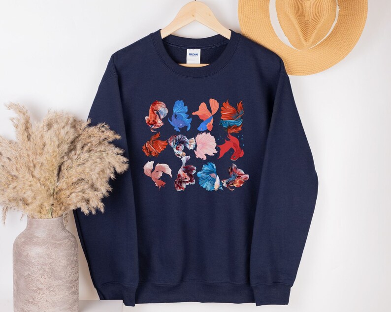 May include: A navy blue sweatshirt with a colorful pattern of betta fish. The fish are arranged in a grid pattern and are in various shades of red, orange, blue, and pink.