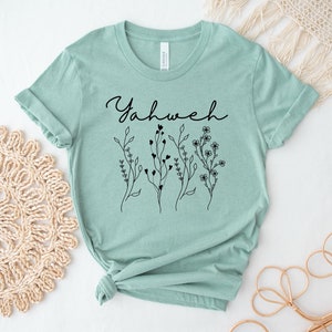 May include: A light green t-shirt with the word "Yahweh" written in black cursive script. The shirt has a floral design with black outlines.