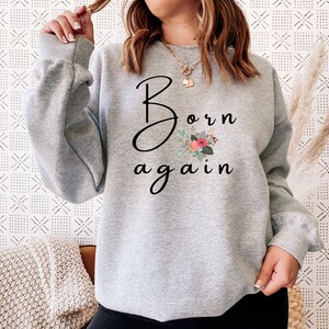 Puede incluir: Sudadera gris con el texto "Born again" en letras cursivas negras y un dise&ntilde;o floral en rosa, rojo y verde.