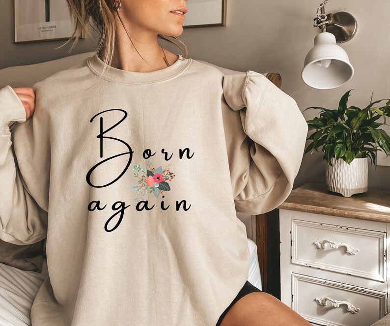 Puede incluir: Una sudadera beige con el texto "Born again" en letra negra con un dise&ntilde;o floral en rosa, rojo y verde.
