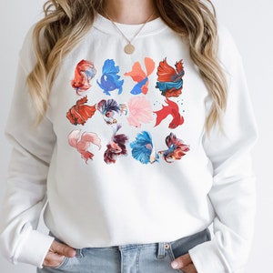 May include: White sweatshirt with a colorful pattern of betta fish. The fish are in various shades of red, blue, pink, and orange.