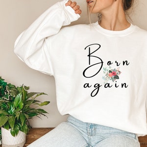 Puede incluir: Sudadera blanca con el texto "Born again" en letra negra con un dise&ntilde;o floral en rosa, rojo y verde.