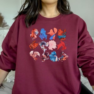 May include: A maroon sweatshirt with a colorful design of betta fish. The fish are in various shades of red, blue, pink, and orange.