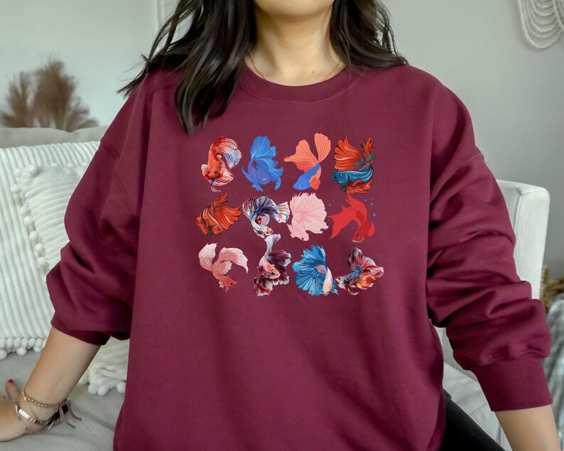 May include: A maroon sweatshirt with a colorful design of betta fish. The fish are in various shades of red, blue, pink, and orange.