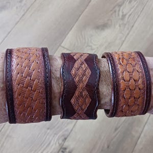 May include: Three brown leather wrist cuffs with embossed designs. The cuffs feature intricate patterns, including a woven texture, diamond shapes, and a geometric design. The leather is a warm brown color with darker brown edges.