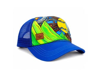 Könnte beinhalten: Eine blaue Trucker-Cap mit einem lebendigen Grafikdruck von hellgrünen Bergen, einer großen gelben Sonne und bunten Sternen vor einem dunklen Himmel. Die Kappe hat einen einfarbig blauen Schirm, ein Netzgewebe auf der Rückseite und ein kleines braunes Patch.