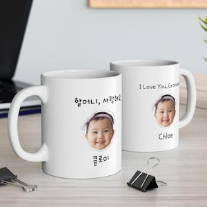 Puede incluir: Dos tazas de cerámica blancas con la foto de una bebé. Una taza tiene texto coreano y el nombre "Chloe". La otra taza dice "I Love You, Grandma" y el nombre "Chloe".