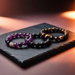 Pode incluir: Três pulseiras de contas sobre uma superfície de ardósia preta. As pulseiras são feitas de ametista, rocha vulcânica e olho de tigre.
