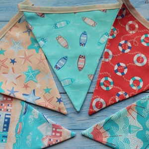 May include: A set of five fabric bunting flags with a nautical theme. The flags are in the shape of triangles and are decorated with different patterns, including surfboards, life preservers, and seashells. The flags are strung together with white string.