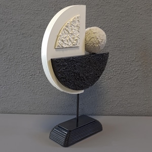 May include: A modern abstract sculpture made of wood and painted in black, white, and gray. The sculpture features a sphere resting on a curved shape, and a base with a layered design.