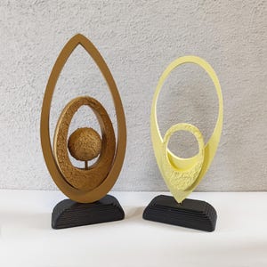 May include: Two abstract sculptures. One is gold with a textured interior and a sphere, set on a black base. The other is yellow, with a similar design, also on a black base. Both are decorative art pieces.