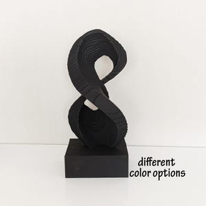 Op de afbeelding: Zwarte abstracte sculptuur met een draaiend, gelaagd ontwerp. Het beeld staat op een zwarte basis. "verschillende kleur opties" staat onder de basis.