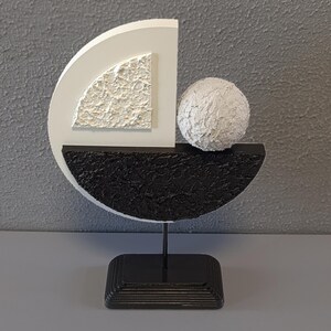 May include: A modern abstract sculpture made of white and black painted wood. The sculpture features a sphere, a half-circle, and a quarter-circle. The sculpture is mounted on a black stand.