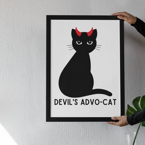 May include: Black and white framed print of a black cat with red devil horns sitting on a white background. The text "DEVIL'S ADVO-CAT" is printed below the cat.