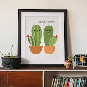 May include: A framed print featuring two cartoon cacti in terracotta pots. The cacti are green with red flowers and smiling faces. The text "Lookin' sharp!" is above the cacti. The frame is black.
