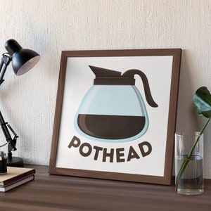 May include: A framed print featuring a cartoon coffee pot filled with dark coffee. The word "POTHEAD" is printed in brown letters below the coffee pot. The frame is brown and the background is white.