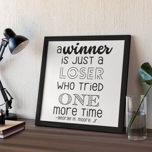 Peut inclure: Citation motivationnelle en noir et blanc dans un cadre noir sur une surface en bois. La citation dit "a winner is just a loser who tried one more time -george m. moore, Jr."