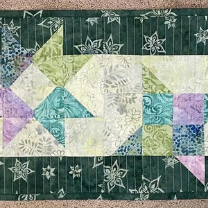 May include: A green table runner with a white border and a row of five patchwork stars in shades of green, purple, and blue. The stars are made of fabric with a floral pattern.