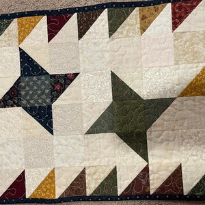 May include: A table runner with a patchwork design featuring a repeating pattern of stars made from different fabric scraps. The runner is made in shades of brown, green, gold, and white.