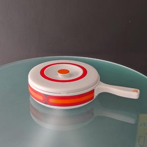 May include: A white ceramic casserole dish with a red and orange geometric design. The dish has a lid with a red circle and a small orange circle in the center.
