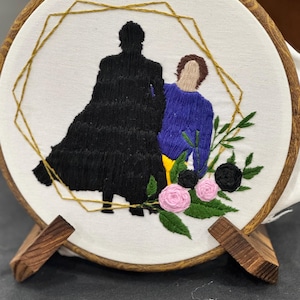 May include: Embroidered hoop art featuring two figures, one in black and one in blue, with floral accents. The design is set within a gold geometric frame on a white background, displayed on a wooden stand.