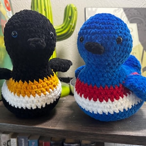 May include: Two round, crocheted bird plushies. The left bird is black with a yellow and white striped belly. The right bird is blue with a red and white striped belly. Both have small black beaks and dark, round eyes, featuring a textured yarn finish.