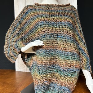 May include: A multi-colored, knitted poncho displayed on a mannequin. The poncho features a mix of brown, blue, yellow, and green hues. The garment has a wide, boat-neck style collar and a loose, flowing fit. The sleeves are short and wide.