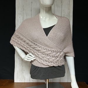 May include: A light brown knitted wrap with a cable knit design across the front. The wrap is displayed on a mannequin, showcasing its draped style. The wrap is made of a soft, textured yarn.