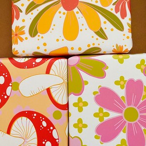 May include: Three different wrapping paper designs featuring a retro style. The first design has large yellow flowers with green leaves on a white background. The second design has red and white mushrooms on a light orange background. The third design has pink flowers with yellow centers and green leaves on a white background.