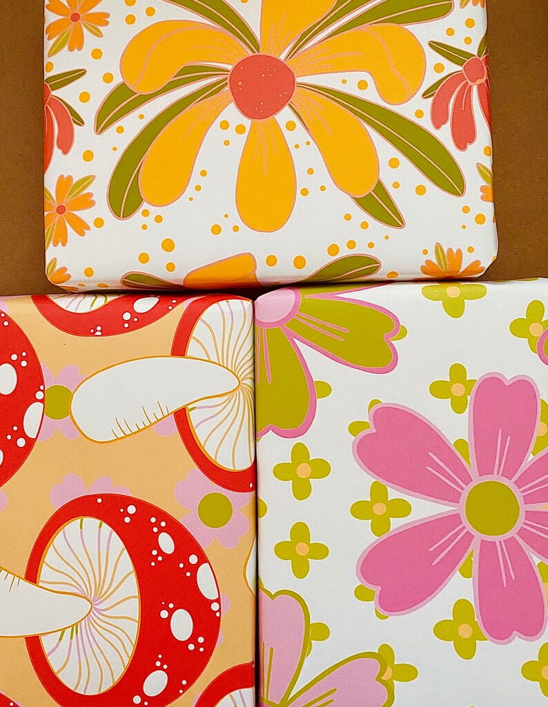May include: Three different wrapping paper designs featuring a retro style. The first design has large yellow flowers with green leaves on a white background. The second design has red and white mushrooms on a light orange background. The third design has pink flowers with yellow centers and green leaves on a white background.