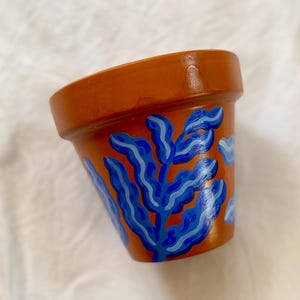 May include: A terracotta-colored ceramic plant pot with a hand-painted design of blue and light blue leafy patterns. The pot has a slightly angled shape and a smooth, glossy finish, suitable for indoor or outdoor use.