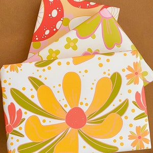 May include: Three sheets of wrapping paper with different floral patterns. The top sheet features a red and white mushroom design, the middle sheet has pink and green flowers, and the bottom sheet has a large yellow flower with green leaves.