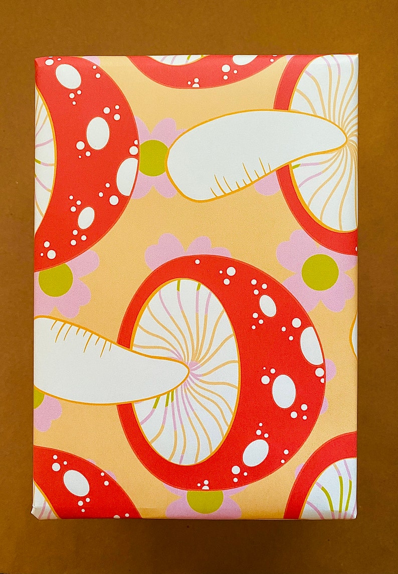 May include: A sheet of wrapping paper with a repeating pattern of red and white mushrooms on a light orange background. The mushrooms have white spots and a white stem.