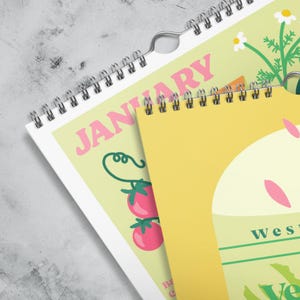 May include: A spiral-bound wall calendar with a light yellow background. The word "JANUARY" is in pink, with illustrations of tomatoes and flowers. The calendar is open to a page with a yellow background and the word "west" in green.