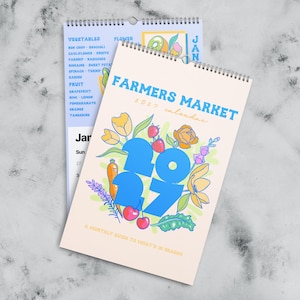 Könnte beinhalten: Ein Farmers Market Wandkalender für 2027 mit Spiralbindung. Das Deckblatt zeigt die großen blauen Zahlen 2027, umgeben von bunten Illustrationen von Blumen, Erdbeeren, Kirschen und einer Karotte auf einem cremefarbenen Hintergrund mit Text.